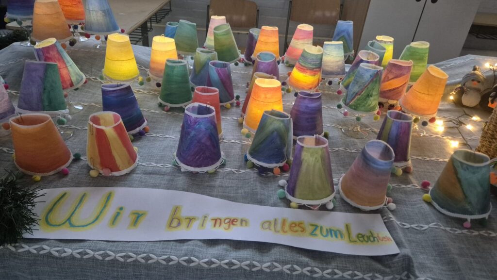 Bunt bemalte Windlichter, davor ein Banner „Wir bringen alles zum Leuchten“