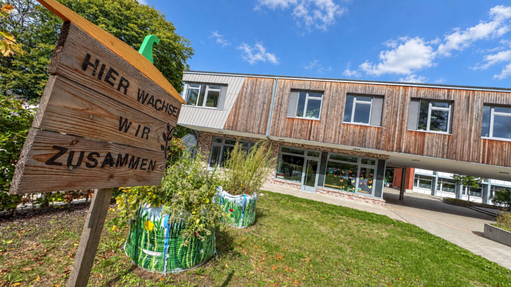 Holzschild „Hier wachsen wir zusammen“ vor dem Schulgebäude