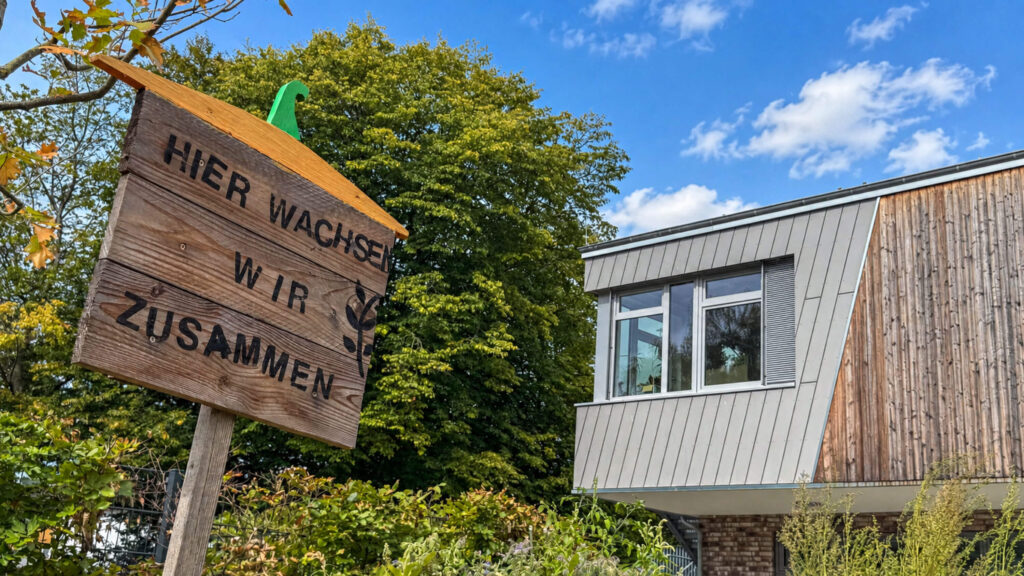 Holzschild „Hier wachsen wir zusammen“ vor einem Gebäudeteil mit Holzfassade