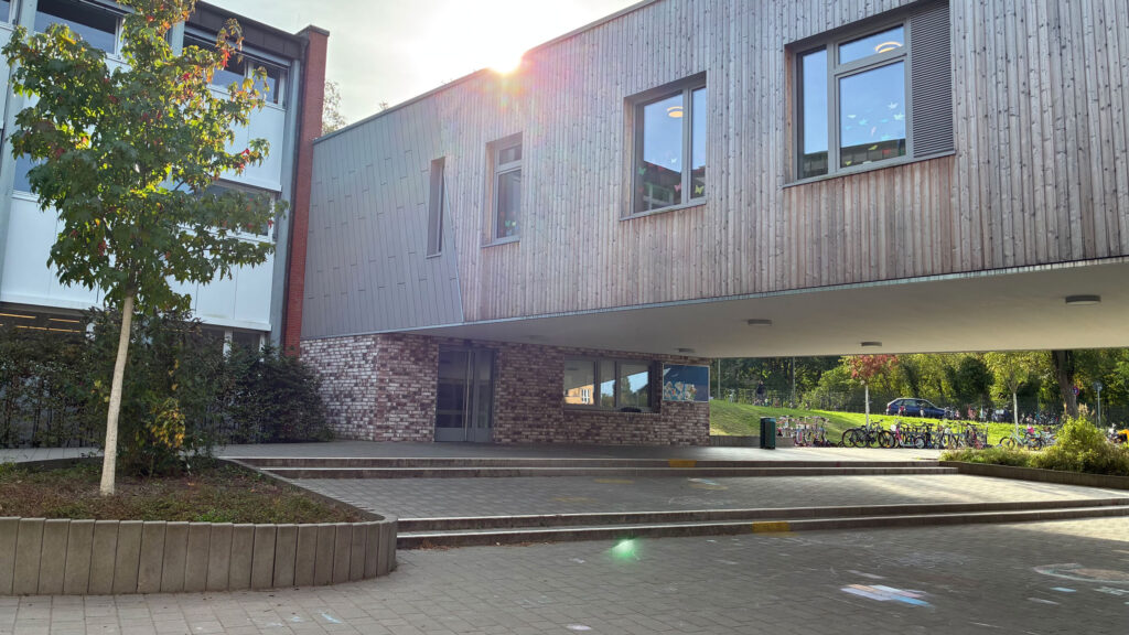 Auskragender Gebäudeteil mit Holzfassade über dem Schulhof