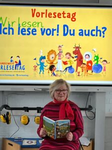 Vorleserin mit Buch vor der Tafel mit dem Plakat „Ich lese vor! Du auch?“